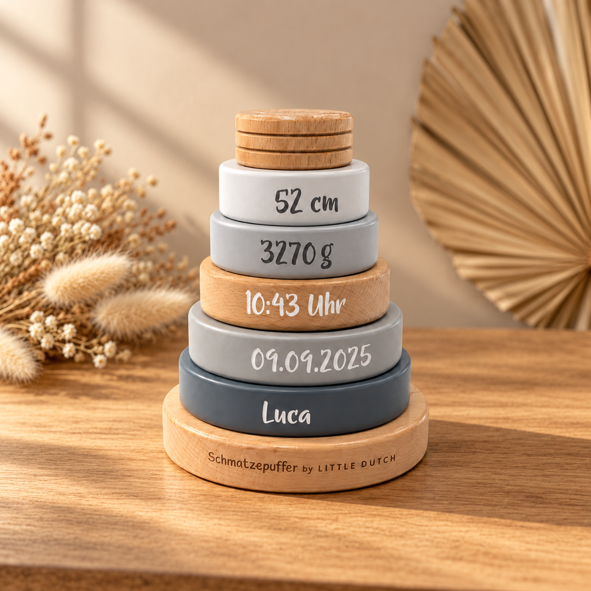Personalisierter Holz-Stapelturm mit Namen Luca und Geburtsdaten in Grau und Naturholz, warm beleuchtet neben Trockenblumen.