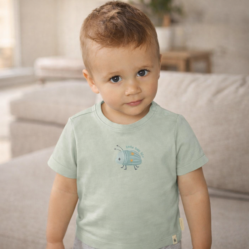 Kleiner Junge trägt ein grünes Kurzarm T Shirt mit Käfer Motiv und steht in einem hellen Wohnzimmer