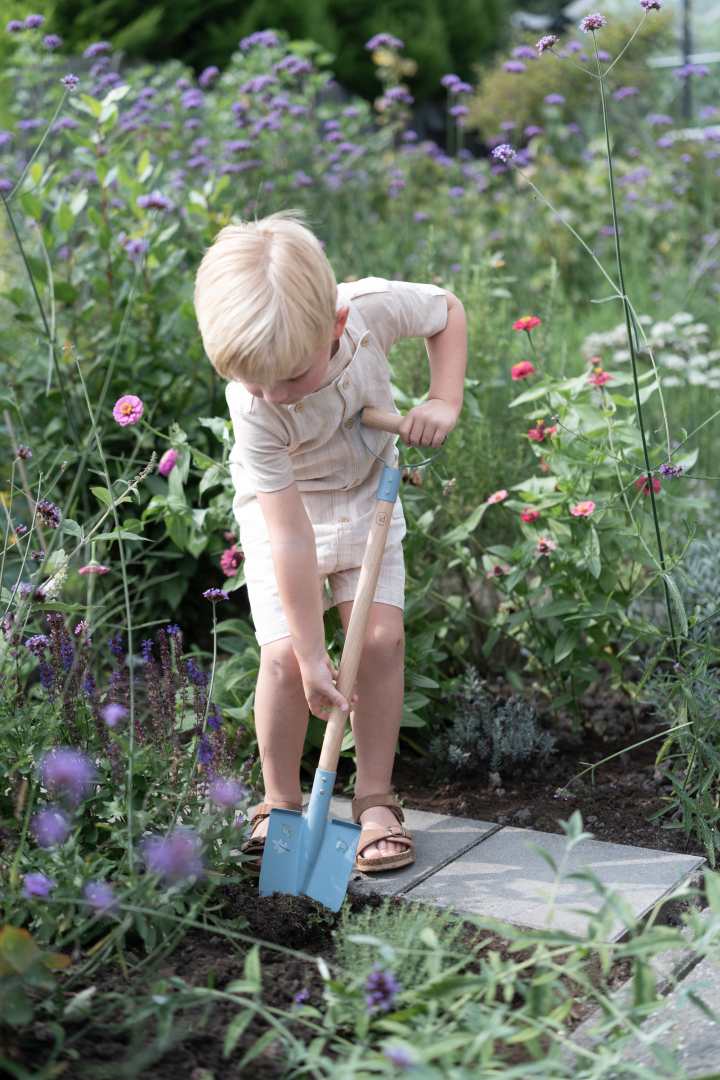 Kind buddelt mit einer blauen Kinder-Gartenschaufel im Blumenbeet. Robuste Gartenschaufel mit Holzstiel für Kinder zum Graben, Pflanzen und spielerischen Entdecken im Garten.