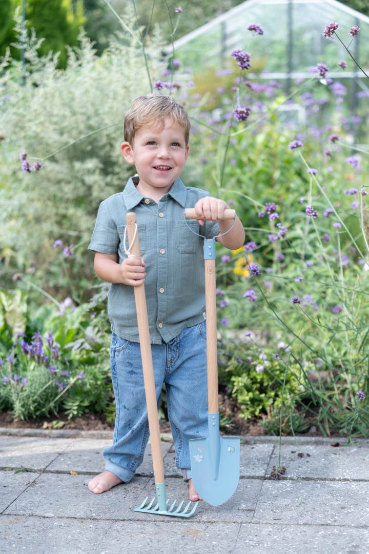 Kind mit Kinder-Gartenschaufel und Kinder-Harke im Garten. Robustes Gartengeräte-Set aus Holz und Metall für kleine Gärtner zum Pflanzen, Buddeln und Spielen im Freien.