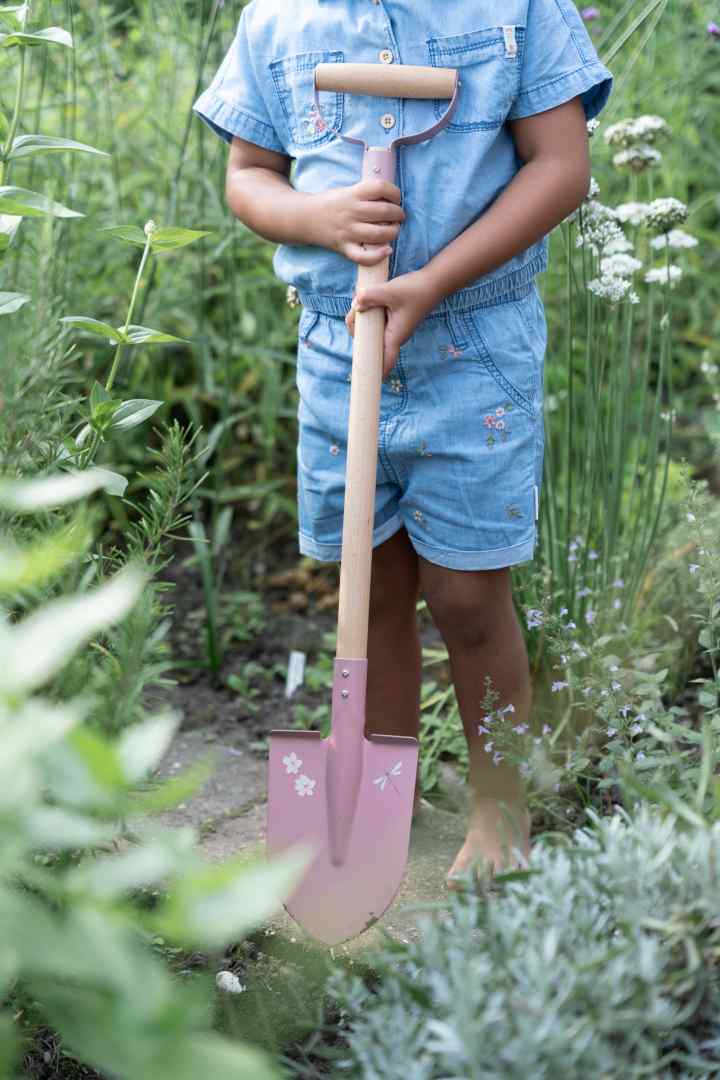 Kind hält eine personalisierte Kinderschaufel in Rosa mit Namen im blühenden Garten. Hochwertige Gartenschaufel für Kinder mit Holzgriff und Metallblatt, personalisiert bei Schmatzepuffer – ideales Geschenk zum Geburtstag, Kindergartenstart oder für kleine Gartenfreunde.